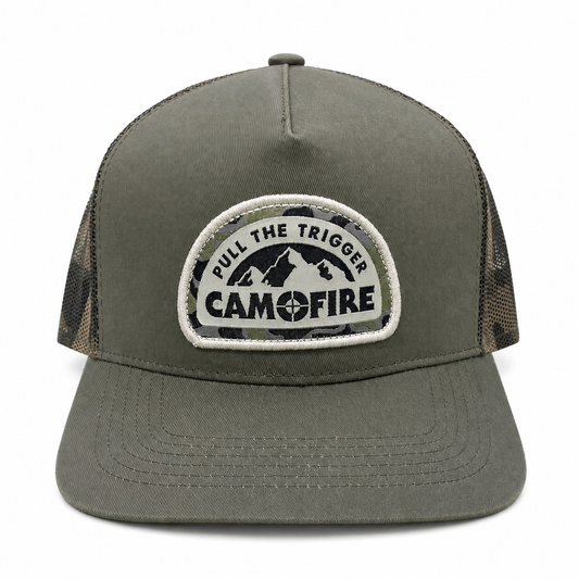 An olive green trucker hat with a camouflage-patterned mesh back and a circular embroidered patch on the front that reads 'PULL THE TRIGGER CAMFIRE' with a mountain graphic.