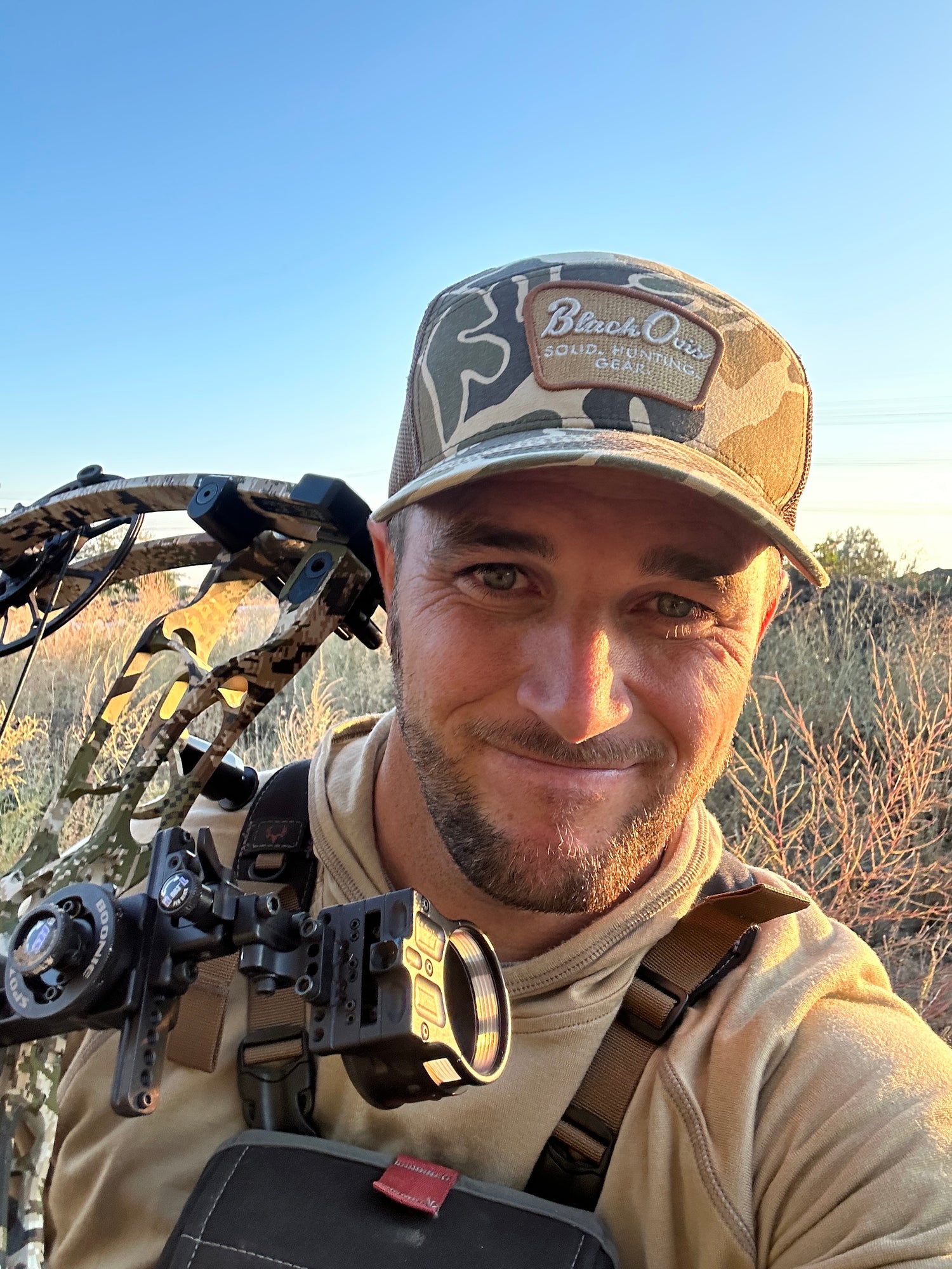A man outdoors wearing a camouflage BlackOvis archery hat, tan hunting shirt, and a compound bow over his shoulder, with a clear blue sky in the background.