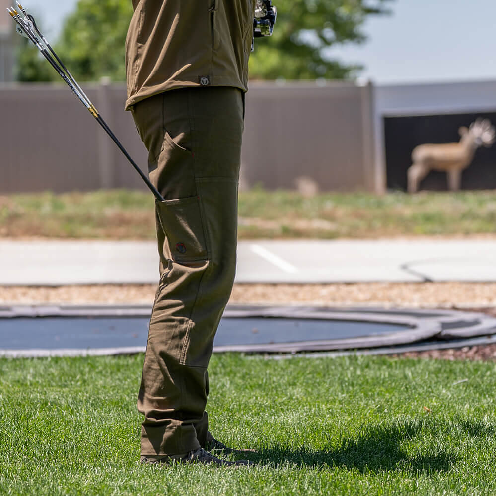 A person standing on grass at an outdoor shooting range, wearing olive green tactical-style pants and a matching olive jacket, holding a bow. A deer target is visible in the background.