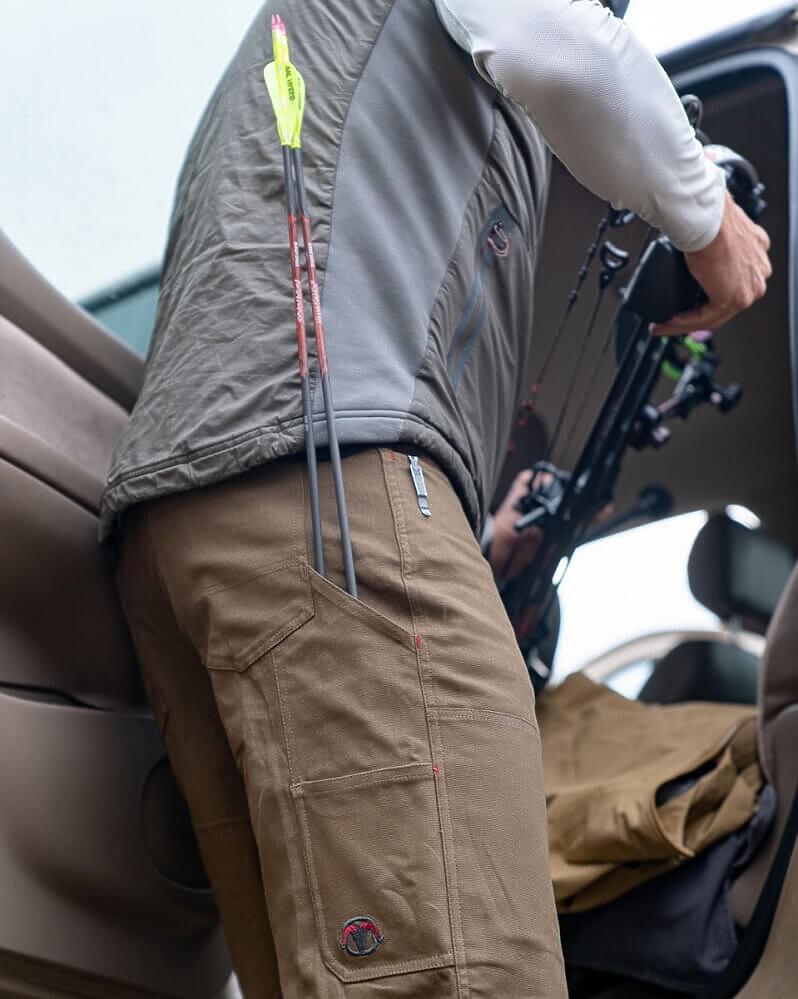A person wearing tan hunting pants with a zippered pocket holding a bow, standing inside a vehicle. The pants feature a small circular logo patch near the lower left leg.