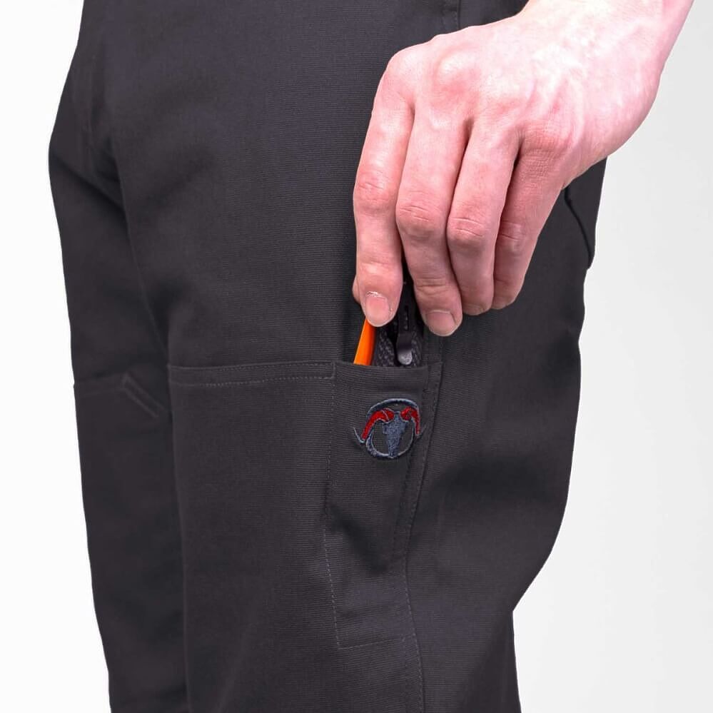Close-up of a person wearing black work pants, showing a hand reaching into a side pocket to pull out a bright orange pencil. A small embroidered logo patch is visible on the pocket.