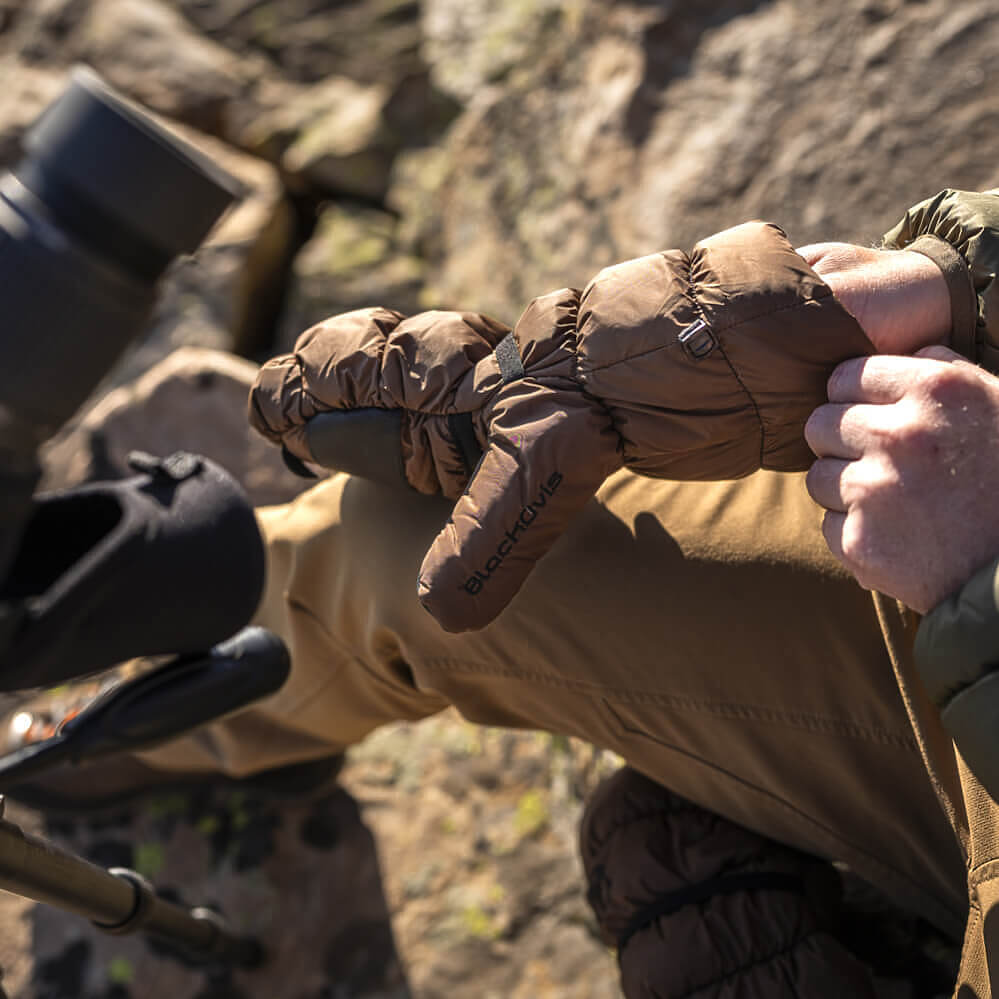 A close-up lifestyle shot of a person wearing a brown insulated puffer glove over a tan winter jacket, sitting outdoors near a spotting scope.
