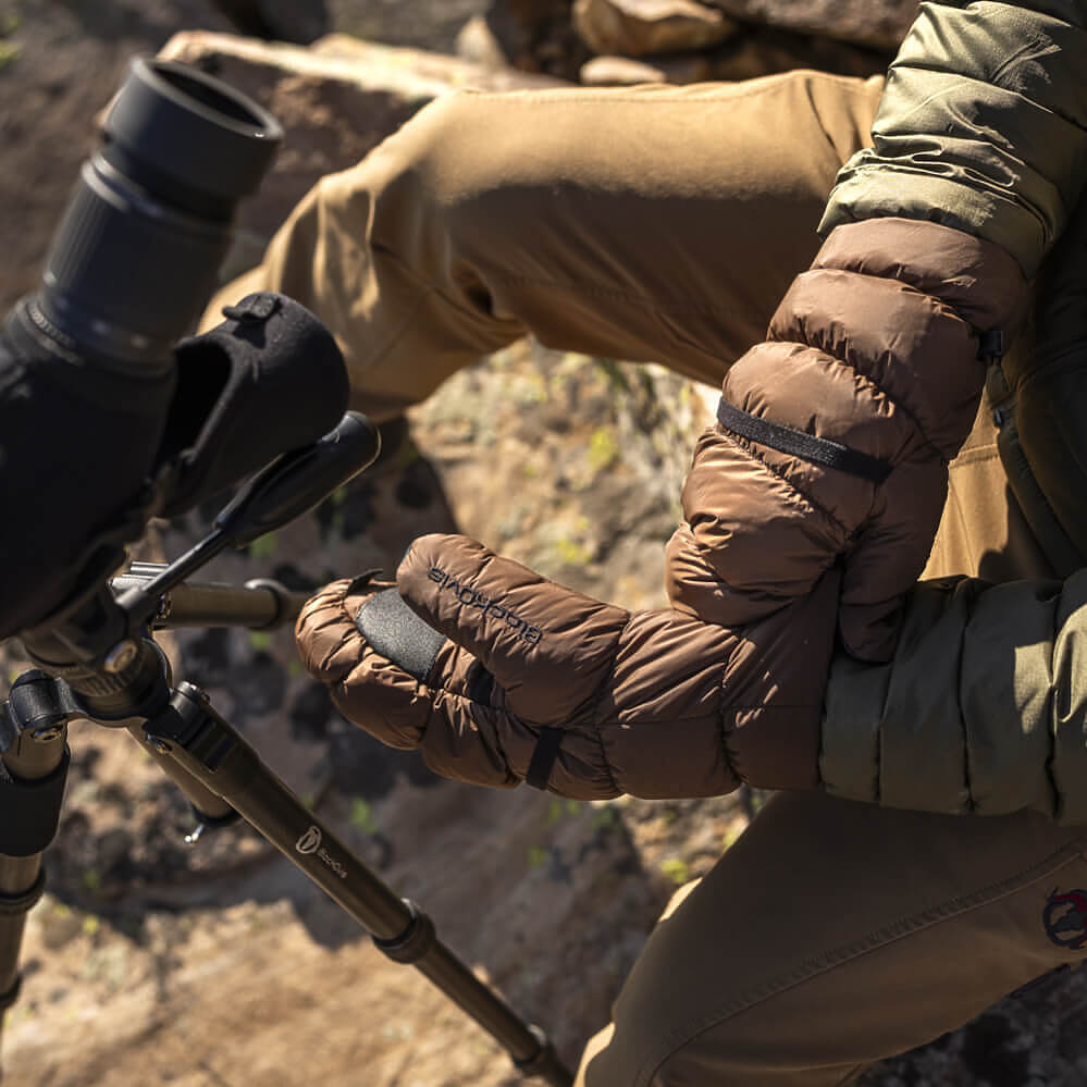 A close-up outdoor shot of a person wearing brown puffer gloves while adjusting a spotting scope on a tripod. The person is also wearing tan pants and a green jacket, with a rocky, natural background.