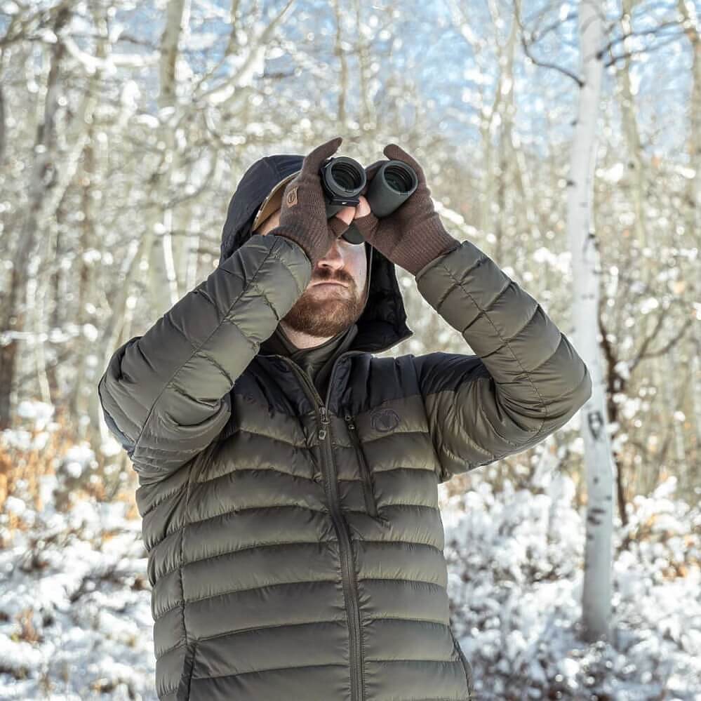 A man wearing a dark olive green puffer jacket and brown gloves looks through binoculars in a snowy forest.