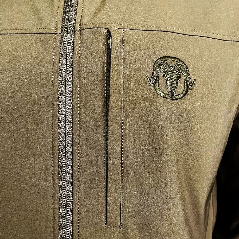 Close-up of a tan technical jacket featuring a vertical zippered chest pocket with a dark embroidered ram skull logo on the left side.