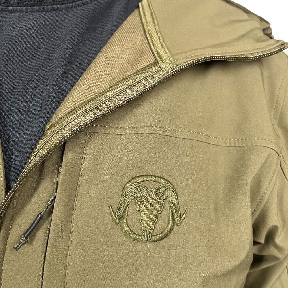 Close-up of a tan outdoor jacket with a tonal embroidered ram skull logo on the chest, shown worn over a dark gray shirt.