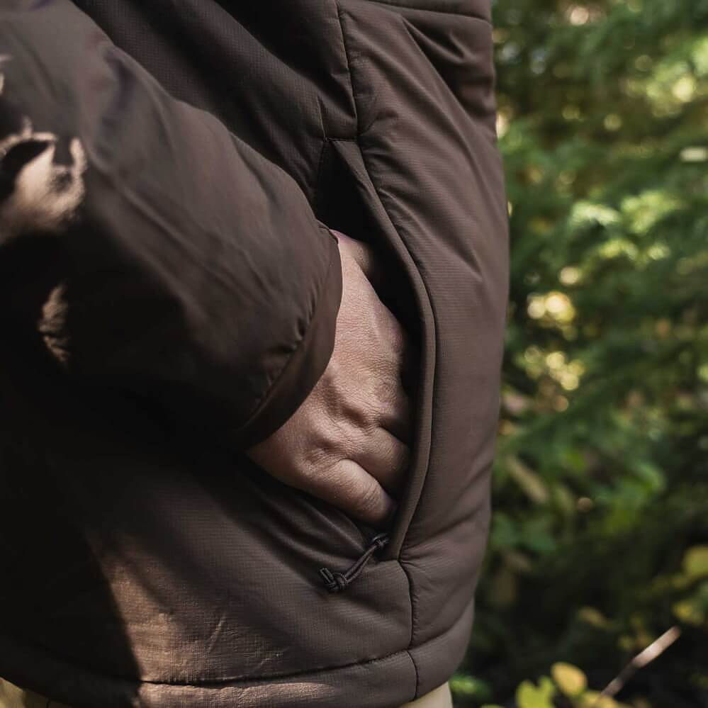 Close-up of a person wearing a dark brown puffer jacket, showing a hand reaching into a side pocket, with a blurred green outdoor background.