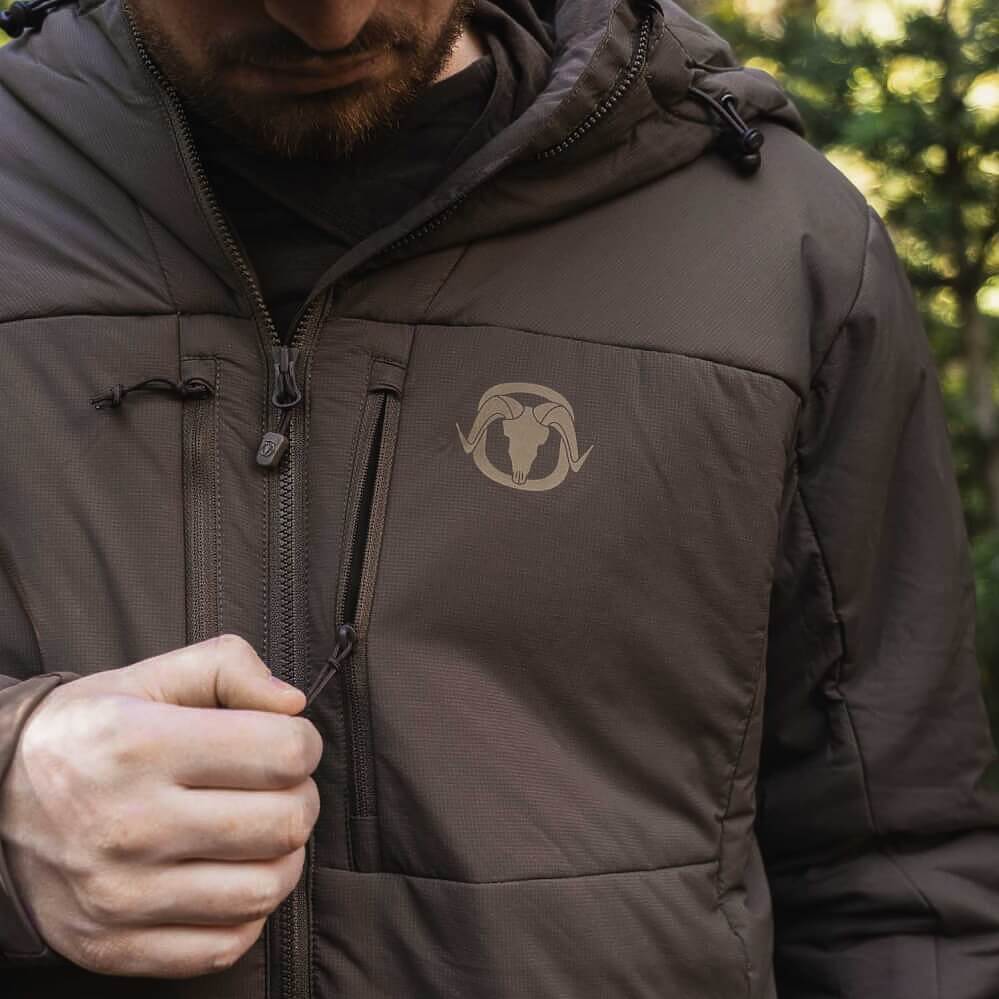 A close-up of a man wearing a dark brown hooded jacket with a subtle light brown ram logo on the left chest, set against a blurred outdoor background.