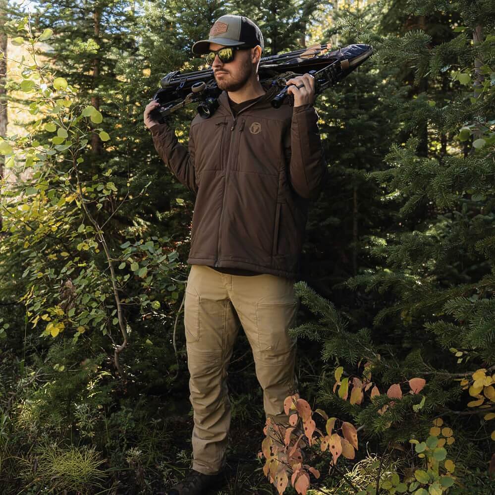 A man wearing a brown zip-up hunting jacket, khaki pants, and a cap stands in a forest holding a crossbow over his shoulders.