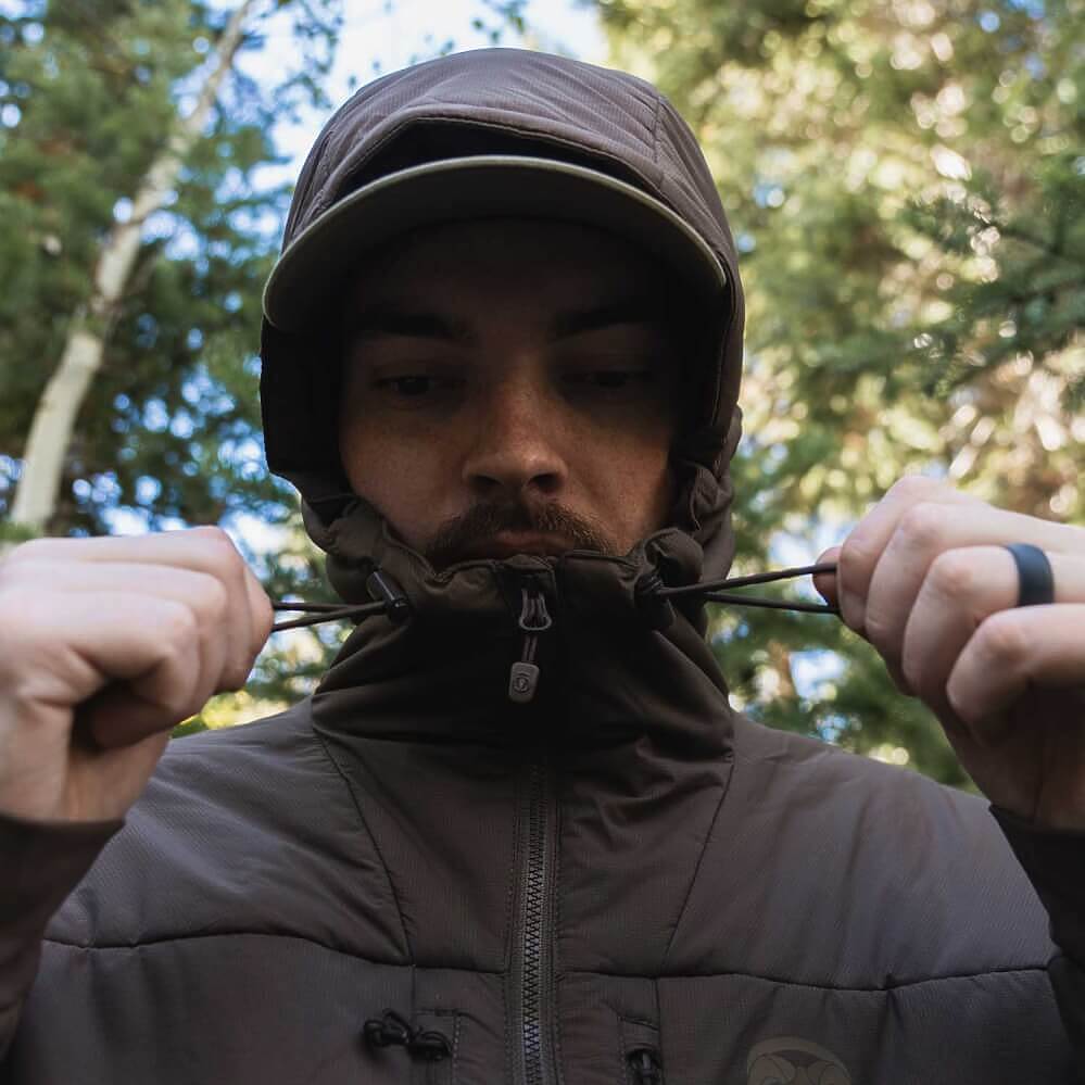 A man wearing a dark brown hooded puffer jacket and matching cap, adjusting the drawstrings of the hood in an outdoor forest setting.