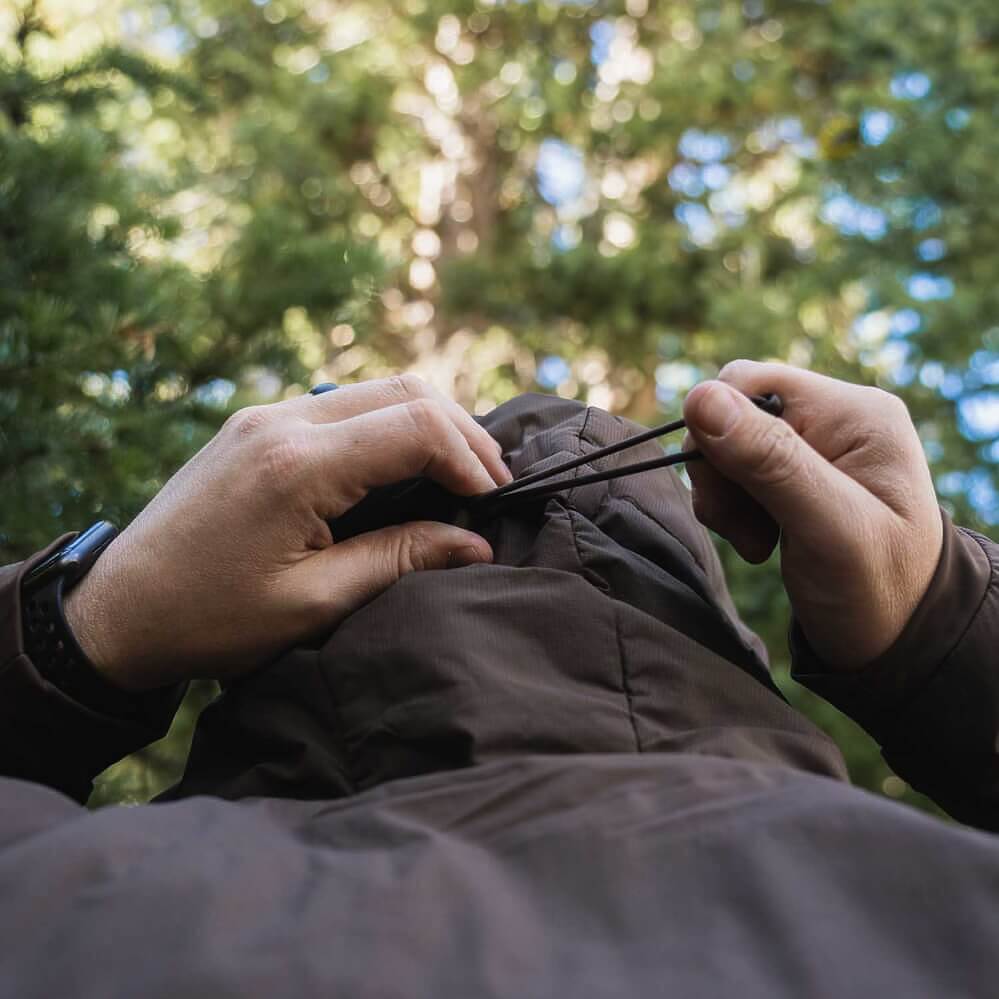Close-up of a person outdoors adjusting the drawstrings of a dark brown puffer jacket, with a blurred forest background.