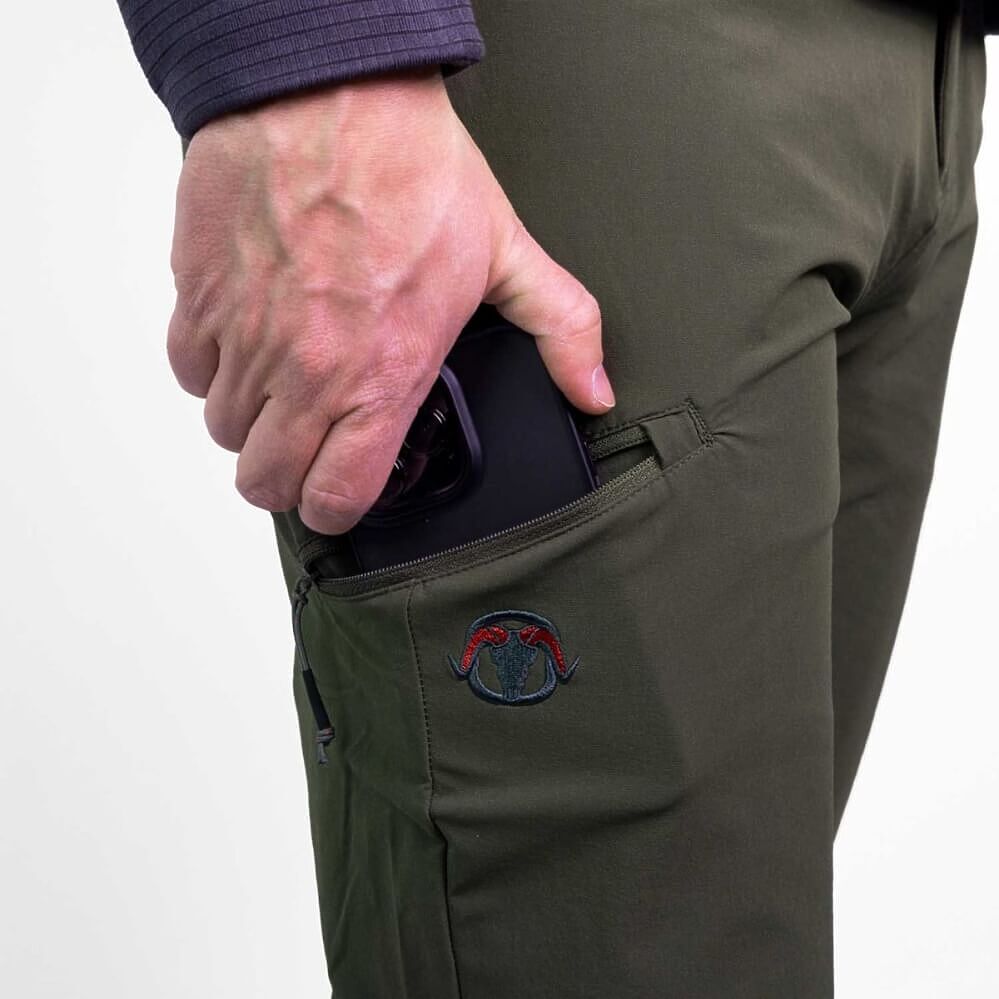 Close-up of a person's hand putting a black smartphone into the side pocket of olive green technical pants, featuring a small embroidered logo patch.