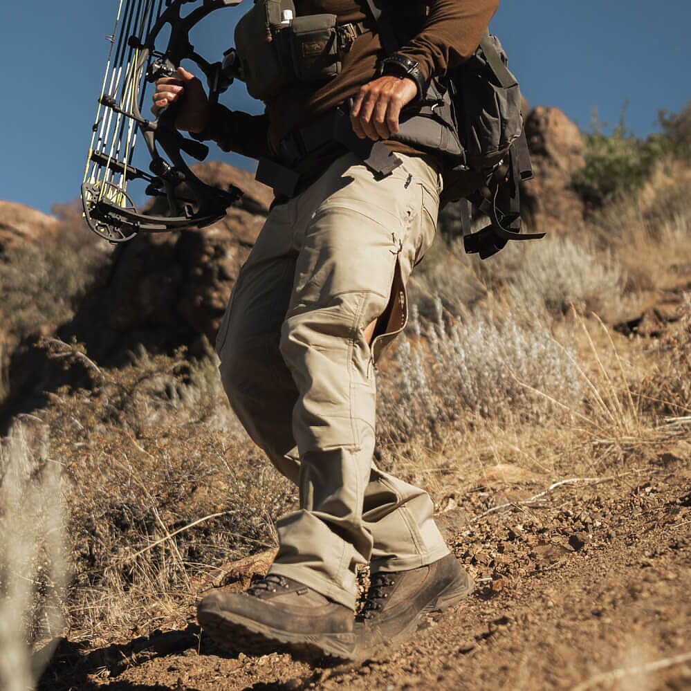 A person hiking on a dry, rocky desert trail wearing khaki cargo pants, a brown jacket, and a black backpack, while holding a compound bow.
