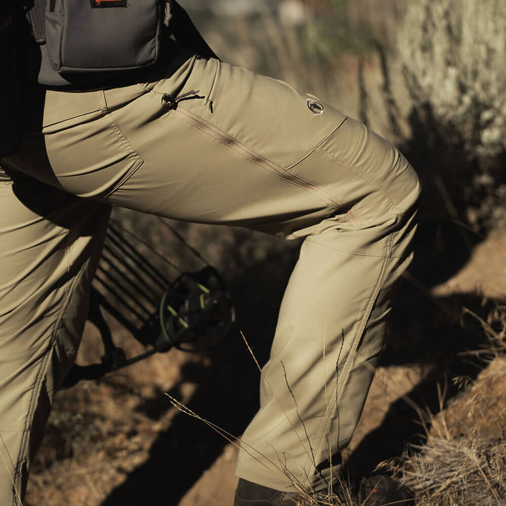 A close-up outdoor shot of a person wearing khaki tactical pants with a zippered pocket and a small logo near the thigh, set against a natural rocky background.