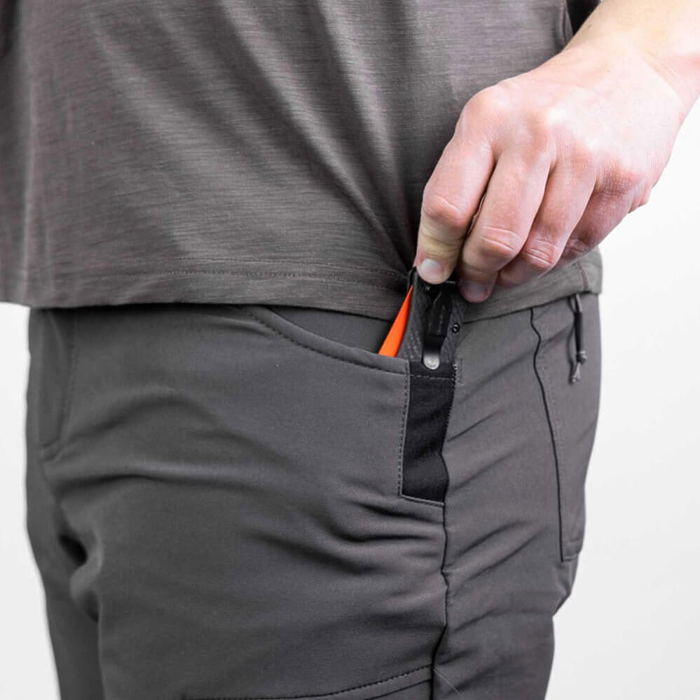 Close-up of a person wearing dark gray technical pants, showing a side pocket with a black zipper and an orange pull tab, with a hand reaching in.