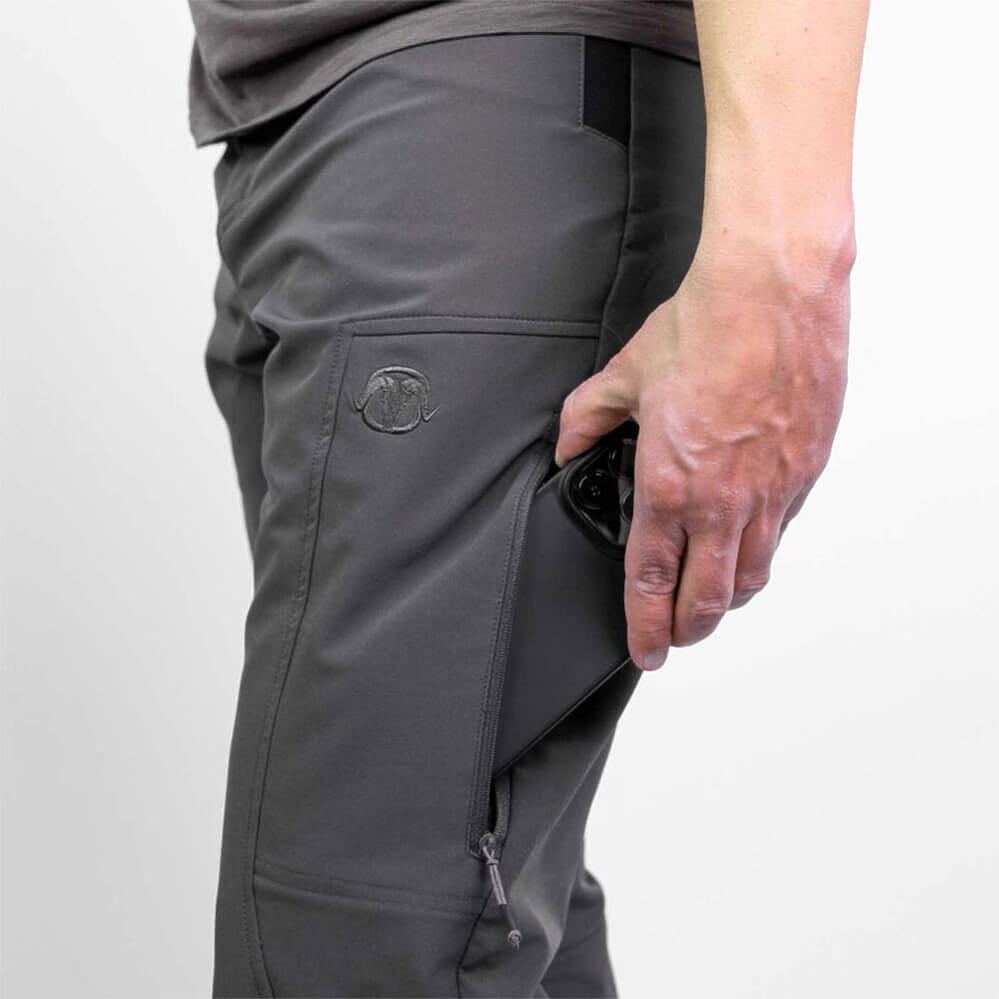Close-up of a person wearing dark gray technical cargo pants, showing a side pocket with a zippered compartment and a small circular logo patch, with a hand reaching into the pocket.